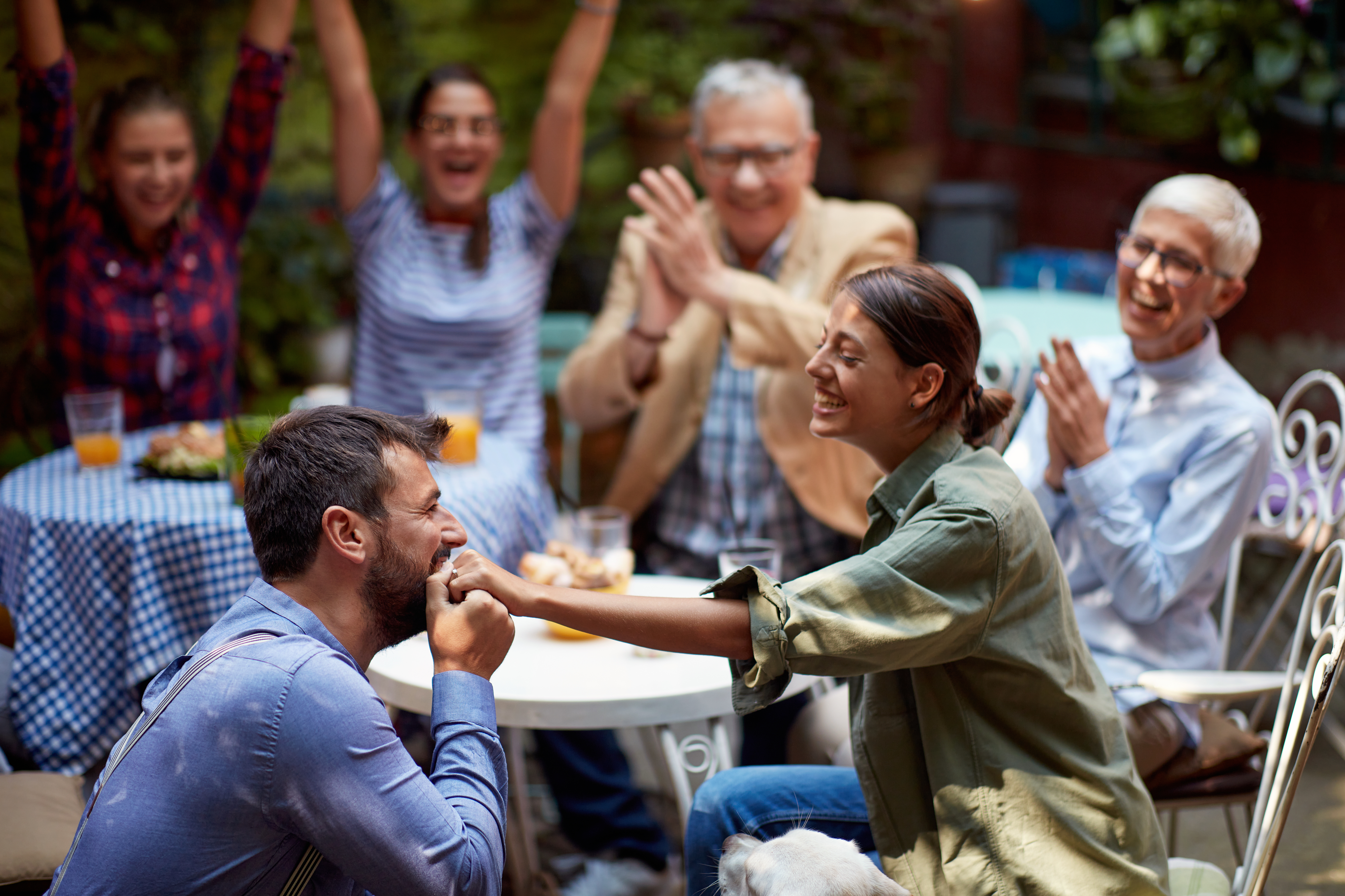 beardy guy kissing the hand of his girlfriend with wedding ring on it, just proposed her in front of closest family and friends, in outdoor cafe
